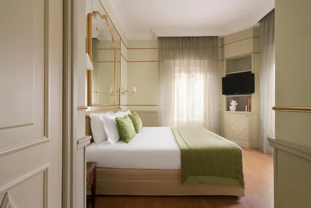 A bedroom with a made double bed with white linen and green cushions, a wall-mounted television, built-in storage, and sheer curtains over a window.