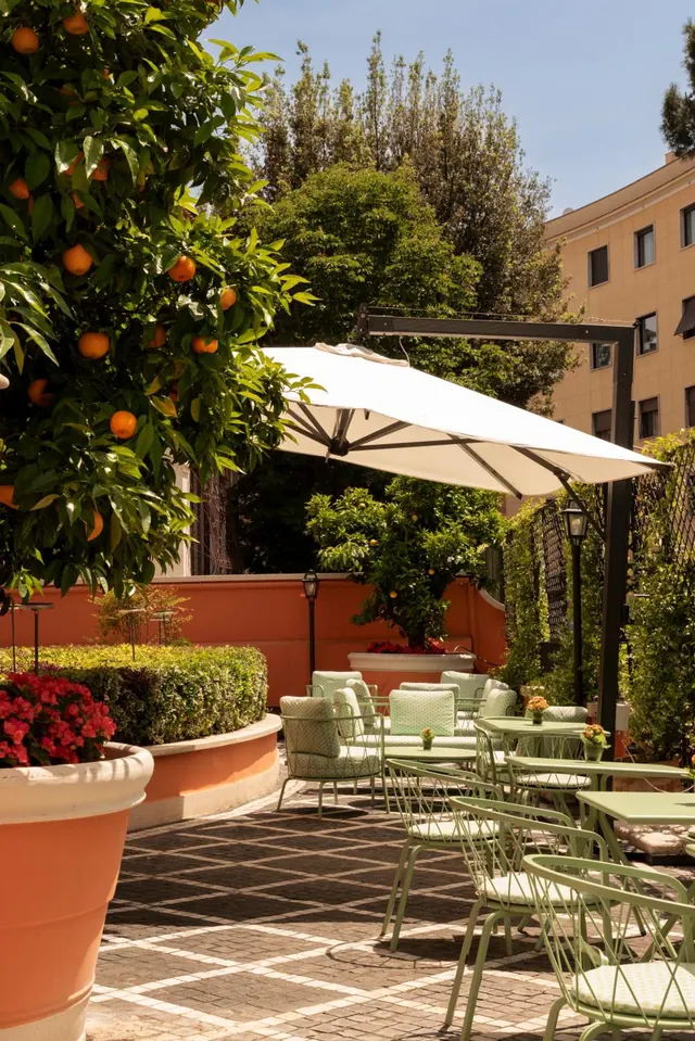 An outdoor courtyard terrace with green metal tables and chairs, a white cantilever umbrella, and an orange tree beside a terracotta wall.