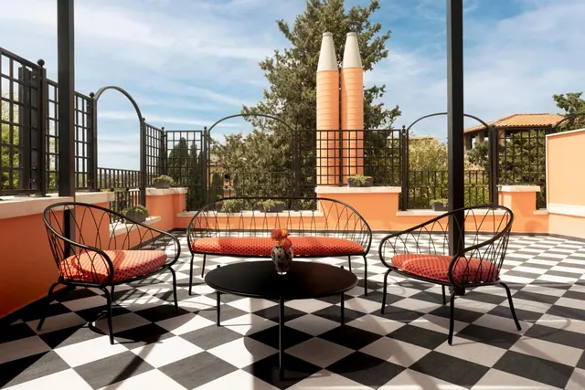 A rooftop terrace with a black and white checkered floor, black metal chairs and a bench with red cushions, a round black table holding a flower vase, orange walls, and tall decorative pillars against a sky with trees behind.