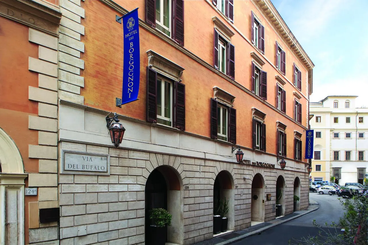 The exterior of Hotel del Borgognoni on Via del Bufalo, with a blue hanging banner, arched ground-floor entrances and a street with parked cars.