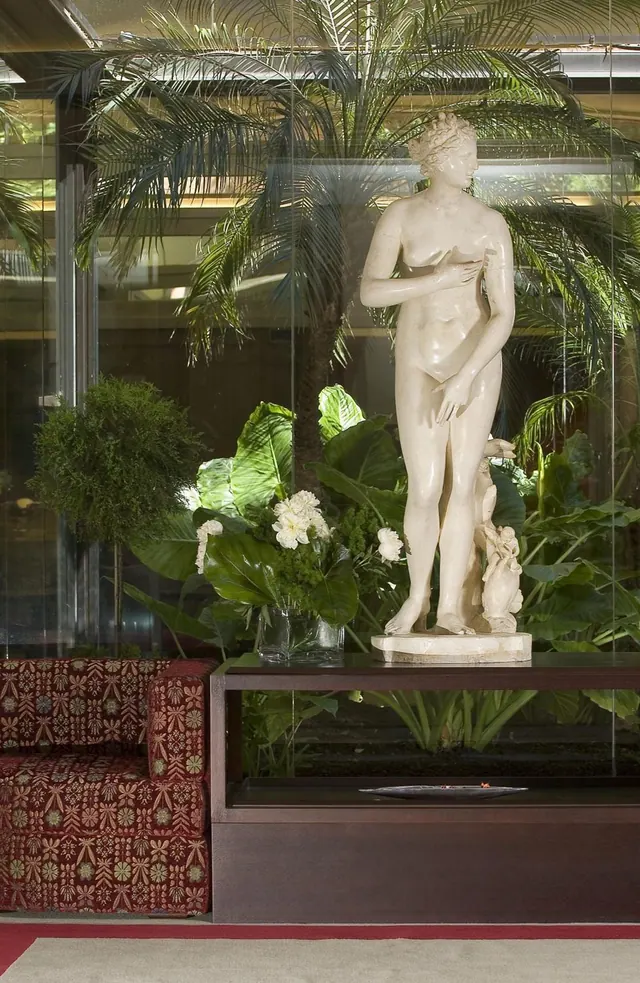 A white marble statue of a standing nude woman sits on a dark display case in a lobby, with palm trees and plants behind glass and a red patterned sofa to the left.