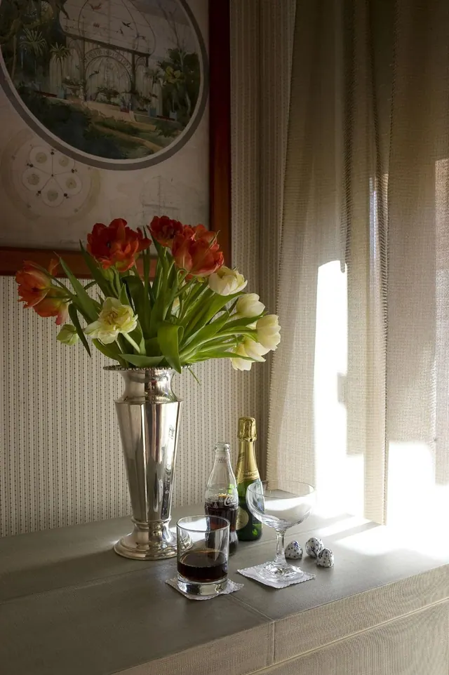 A silver vase of tulips, a champagne bottle, two glasses and small speckled eggs sit on a window ledge beside sheer curtains and a framed circular painting.