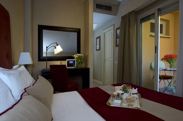 A hotel bedroom with a made double bed and a tea tray on the covers, a desk with a chair, and sliding doors opening onto a balcony with a table and chairs.