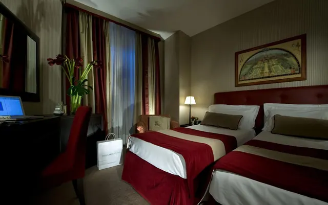 A hotel bedroom with two twin beds under red and cream runners, a desk with a laptop, a red chair, and a shopping bag on the floor.