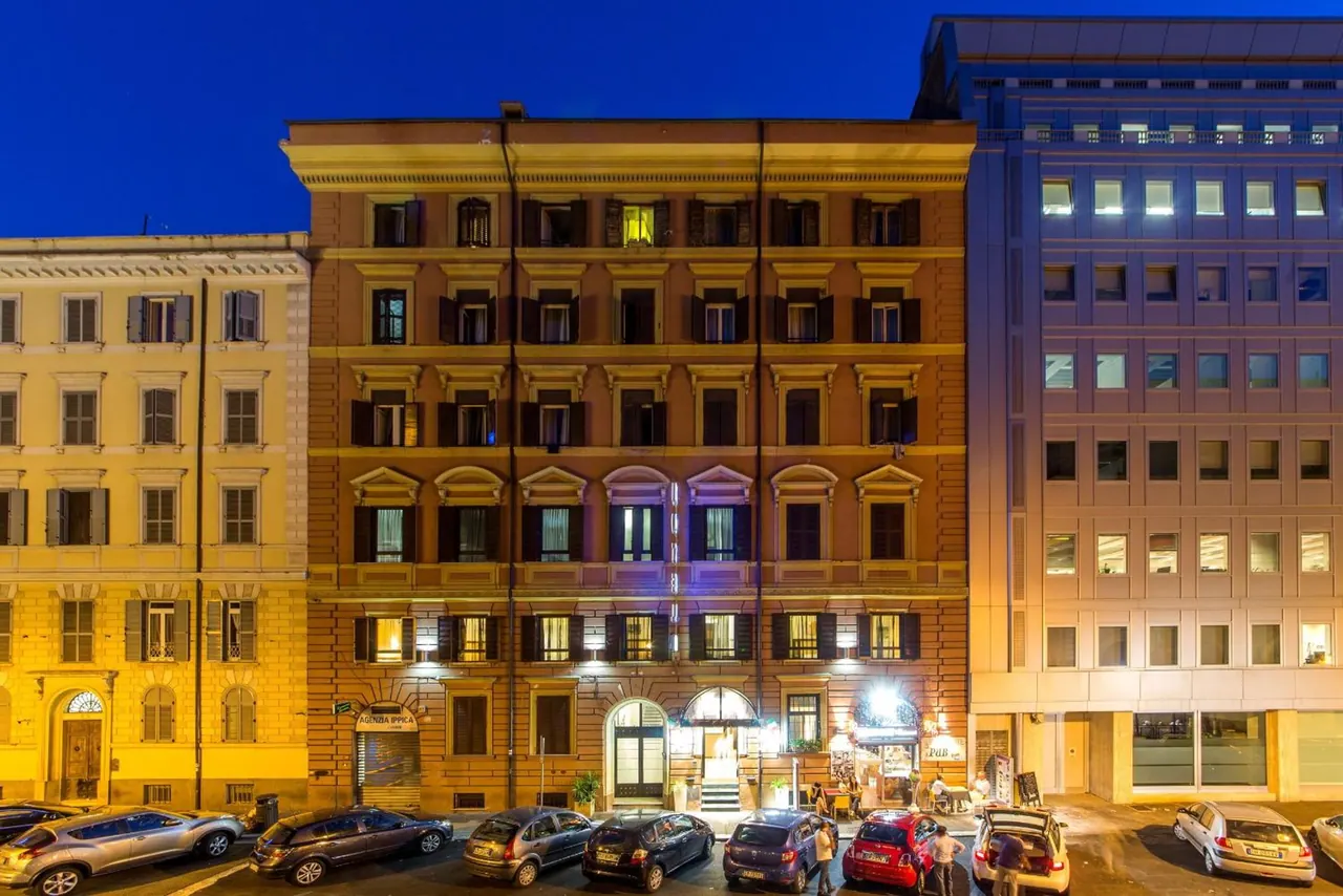 Evening exterior of a multi-storey ochre hotel building with an arched entrance, flanked by older yellow and modern office buildings, with cars parked along the street.