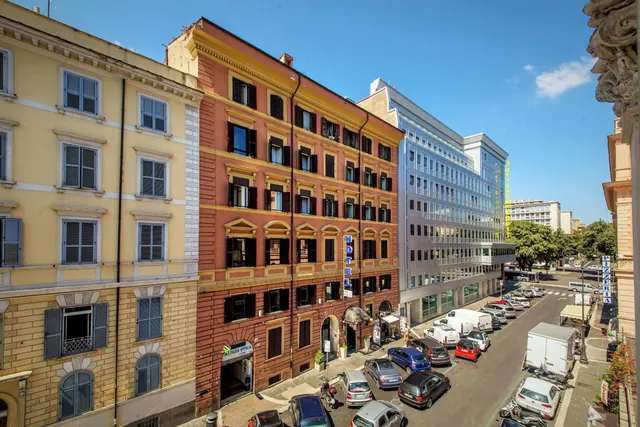 A sunny view down a Rome street from an upper floor, showing a red hotel building with a vertical HOTEL sign, parked cars, vans and scooters.