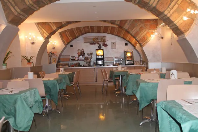 A breakfast dining room with brick vaulted arches, rows of tables with green tablecloths, and a buffet counter with coffee machines at the back.