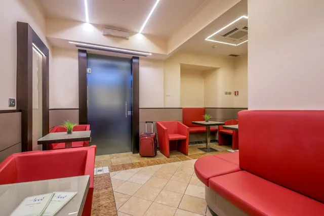 A seating area with red leather sofas and armchairs, small tables, a red suitcase by a frosted glass door, and ceiling light strips.