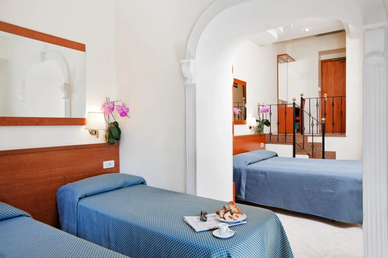 Twin beds with blue bedspreads sit in a room with an arched doorway, and a tray with bread and coffee rests on one bed.
