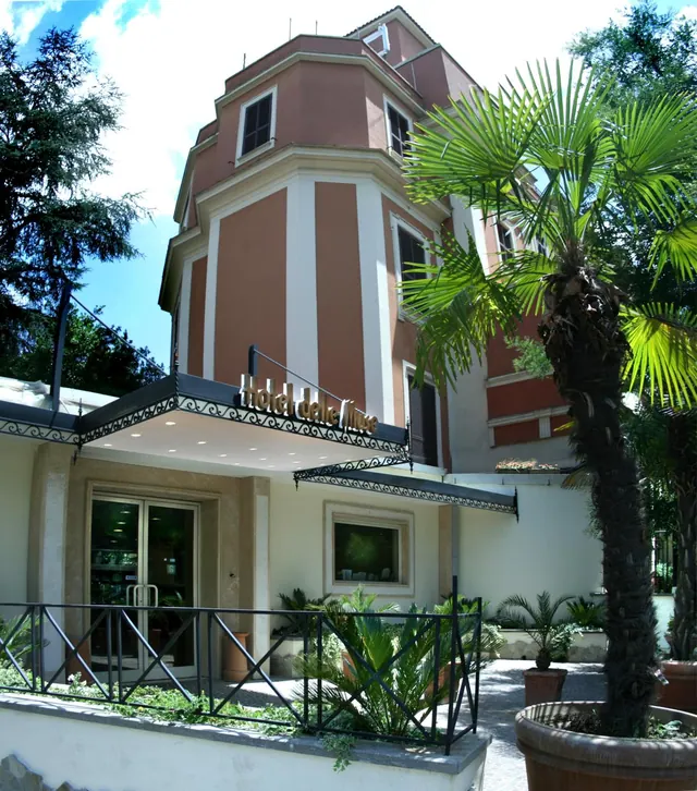 The exterior of Hotel delle Muse shows a multi-storey pink and cream building with a tower, a black canopy over the entrance, and a palm tree in the foreground.