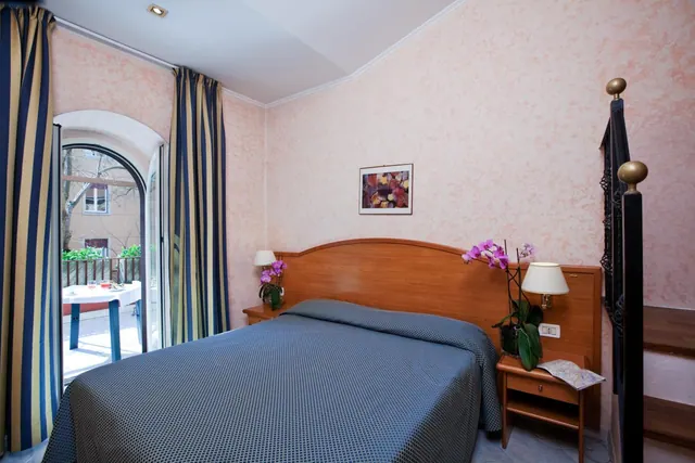 A double bed with a blue patterned cover sits beside a wooden headboard, with an arched door opening onto a terrace with a table and chairs.