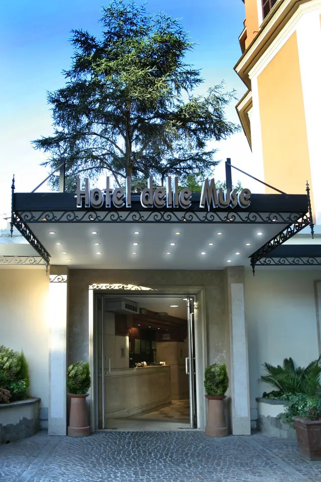 The entrance of Hotel delle Muse, with a lit canopy sign, open glass doors onto a reception desk, potted plants and a cobbled forecourt, with a tree behind the canopy.