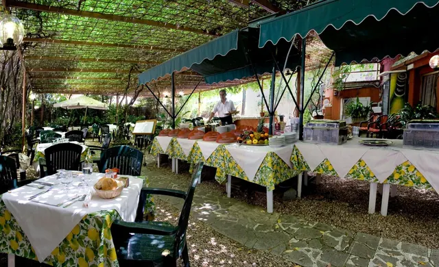 An outdoor restaurant terrace under a vine pergola and green awnings, with lemon-patterned tablecloths, a fruit display and a waiter at the buffet counter.