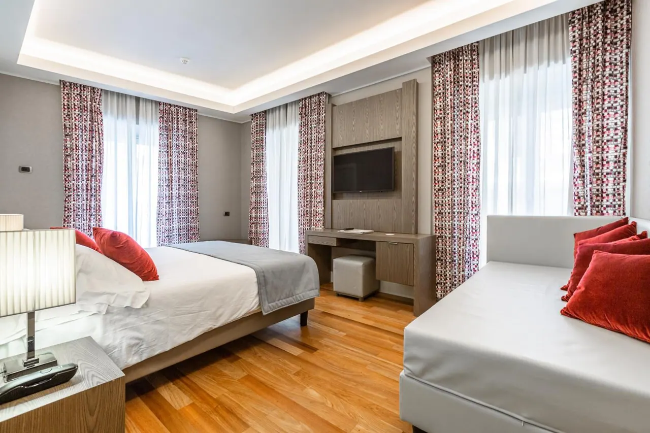 A bedroom with a double bed, a white sofa bed with red cushions, a wooden desk below a wall-mounted television, and tall windows with patterned curtains.