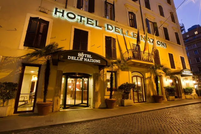The illuminated facade of Hotel Delle Nazioni at dusk, with an arched revolving entrance, potted palms and a cobbled street in front.