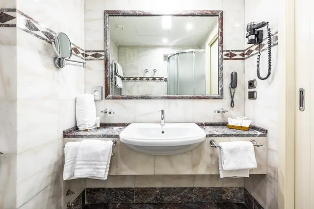 A marble-tiled bathroom with a wall-mounted magnifying mirror, a white basin on a stone countertop, folded towels, a curved glass shower enclosure, and a wall-mounted hair dryer and telephone.