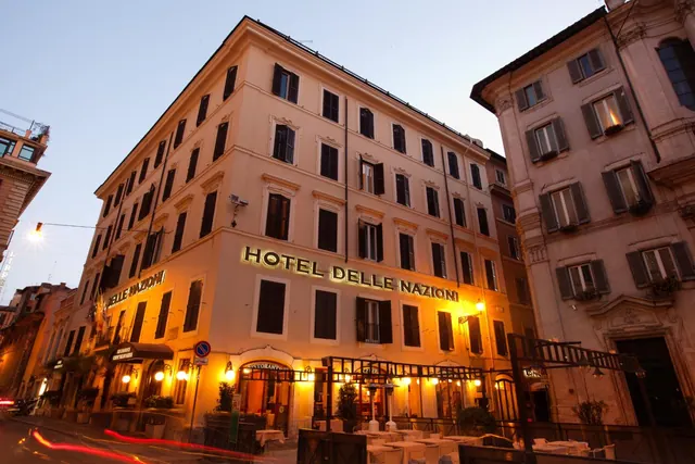 Evening exterior of the Hotel Delle Nazioni building, with its illuminated sign and outdoor dining tables at street level.