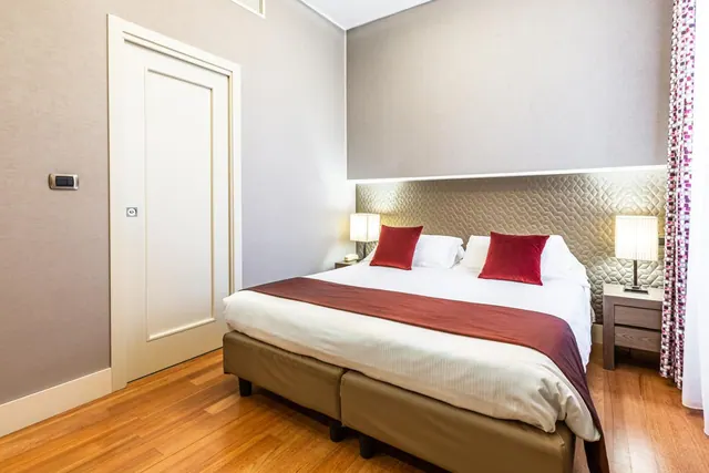 A bedroom with a double bed, red cushions, a quilted headboard, two bedside lamps and a wooden floor.