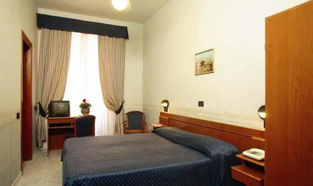 A bedroom with a double bed under a navy bedspread, a wooden wardrobe, a desk with an old television, and an armchair beside curtained windows.