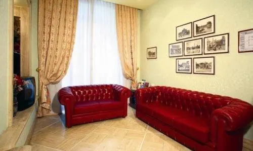 A living room with two red leather tufted Chesterfield sofas, framed prints on the wall, and a window with sheer and patterned curtains.