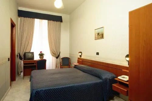 A double bed with a navy bedspread and a wooden headboard sits in a room with a desk holding a small television, a chair, and a wooden wardrobe on the right.