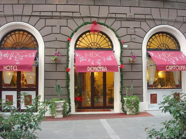 The front of a stone building shows three arched entrances with pink awnings reading HDC, Hotel Demetra and Capitolina, plus green garland and red decorations around the central doorway.