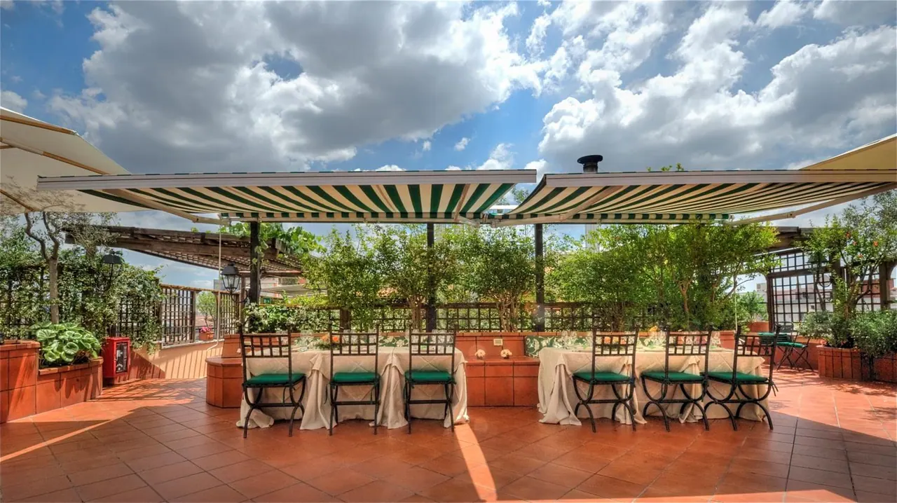 A rooftop terrace with green and white striped awnings, set outdoor tables with white cloths and green-cushioned chairs, and potted trees under a cloudy sky.