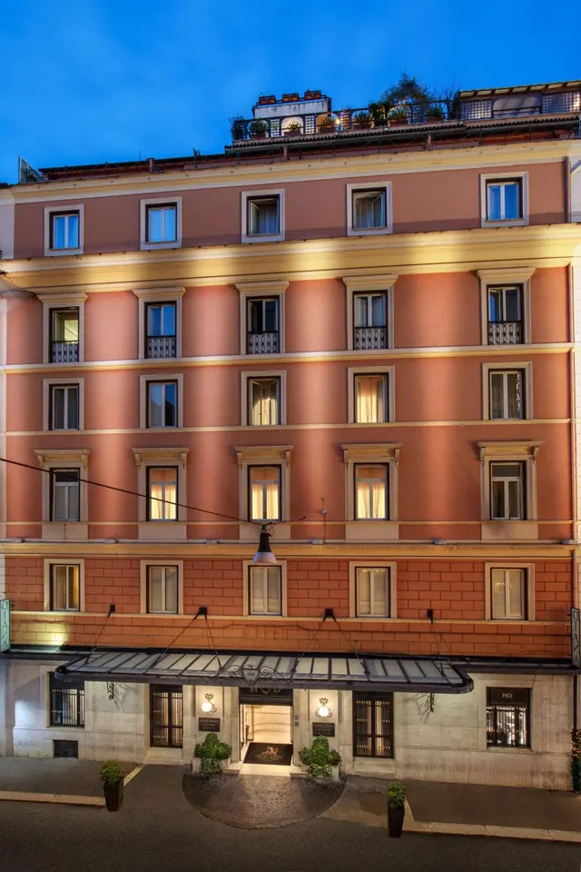 The terracotta facade of a multi-storey hotel building at dusk, with lit windows, a canopy over the entrance and planters at street level.