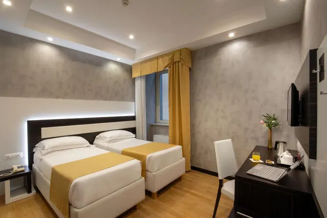 A twin-bedded hotel room with a wooden desk, laptop, kettle tray and wall-mounted television beside the window.