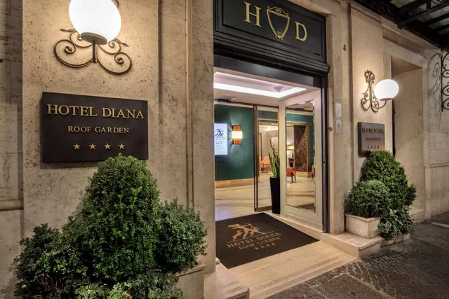 Night view of the hotel entrance with a dark doormat, illuminated wall lamps, a Hotel Diana Roof Garden sign, and trimmed shrubs beside the door.