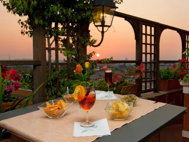 A set table with drinks and snack bowls on a rooftop terrace, with a city view and a distant dome at sunset.
