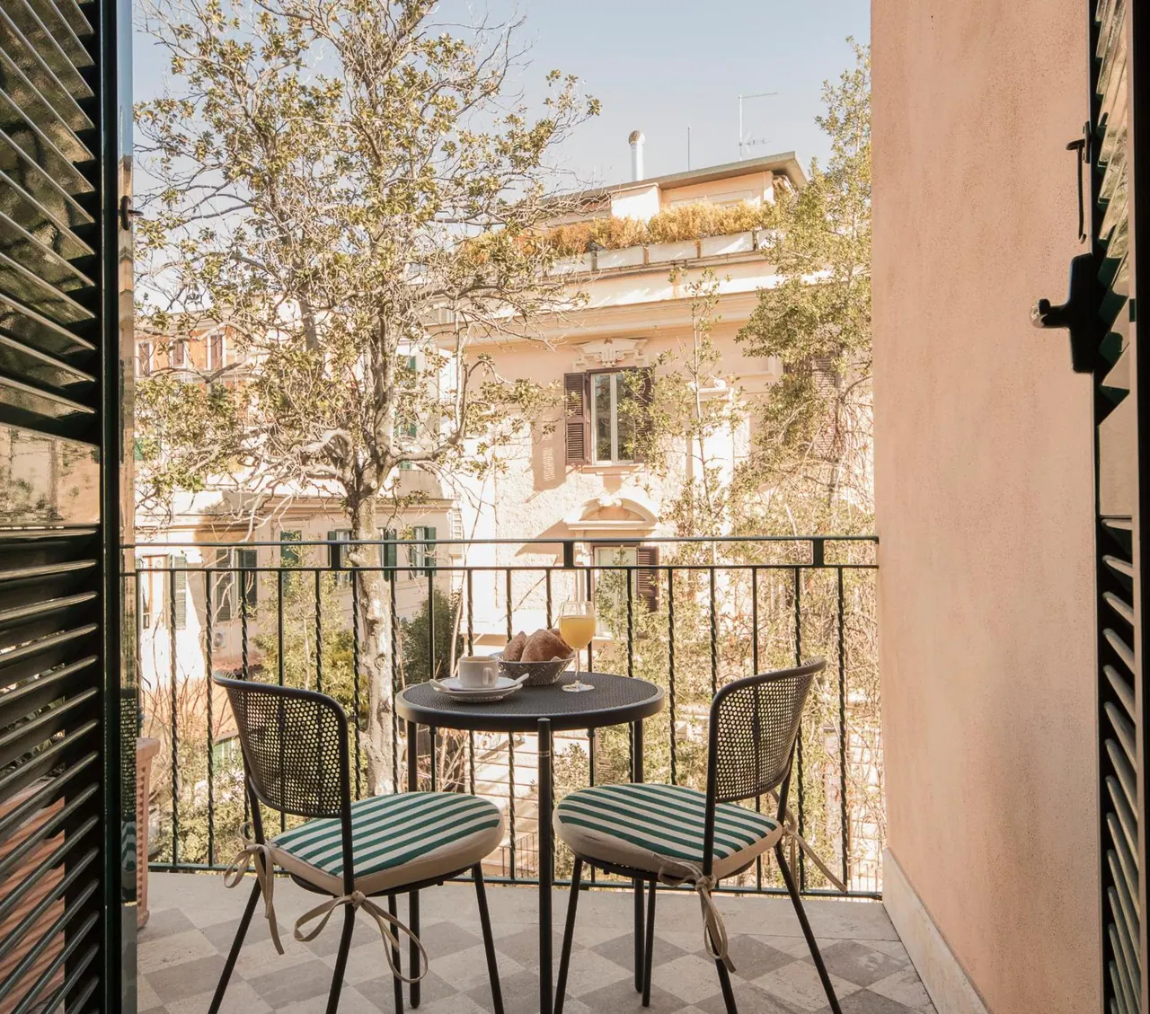 A small round table with two striped-cushion chairs, a coffee cup, croissants and a glass of orange juice sits on a tiled balcony overlooking neighbouring buildings and trees.