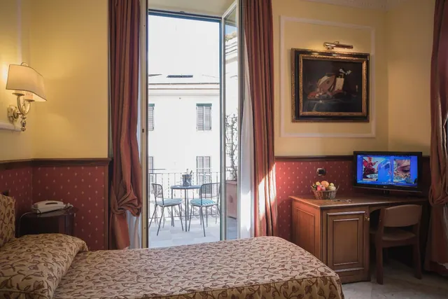A hotel bedroom with a double bed, a desk with a television, and open French doors onto a small balcony with a table and two chairs.