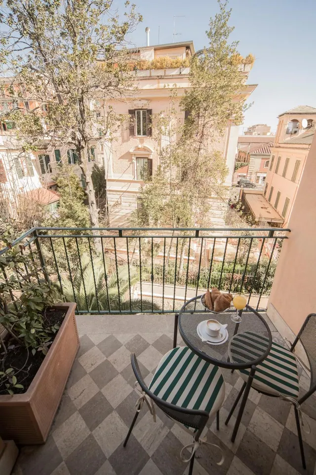 A private balcony with a round metal table, two striped chairs, a coffee cup and a croissant, overlooking neighbouring buildings and trees.