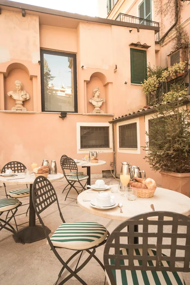 An outdoor courtyard terrace with round stone-topped tables, green-striped chairs, breakfast drinks and bread, and classical busts set in a pink wall.