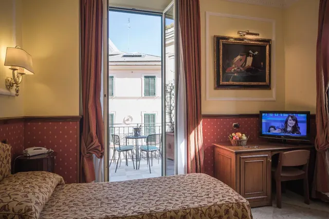 A hotel bedroom with a double bed, a wooden desk with a television and fruit basket, and open French doors onto a small balcony with a table and two chairs.