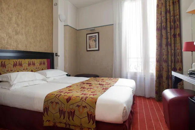 A bright hotel bedroom has a double bed with patterned runner, a desk, seating, and tall curtained windows.