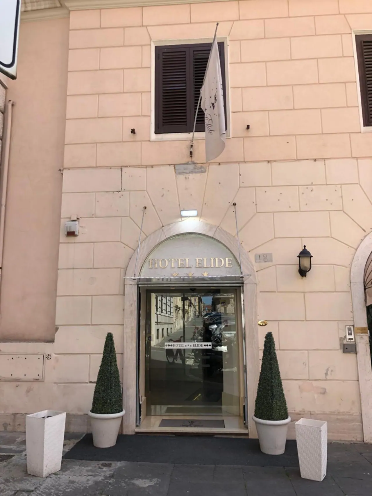 The hotel entrance has a glass door under an arched stone frame with the name Hotel Elide above it, a flag on the facade, and two topiary planters on the pavement.