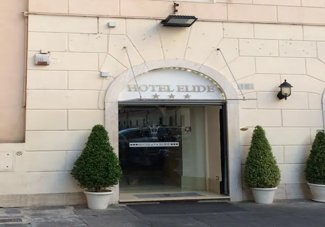 Street-level entrance to Hotel Elide, with an arched stone doorway, a sign reading HOTEL ELIDE with three stars, and three potted topiary shrubs on the pavement.
