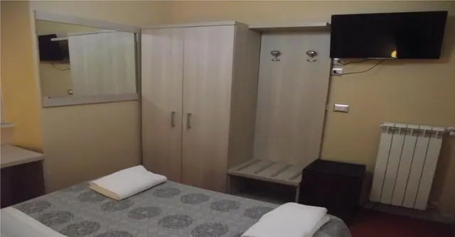 A bedroom with a double bed covered in a patterned grey spread and two folded white towels, a wardrobe, a wall-mounted flat-screen TV and a wall radiator.