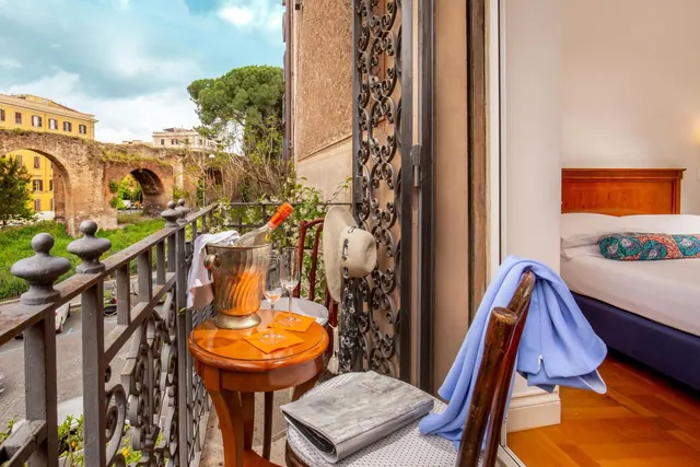 A small wooden table with a champagne bucket, two glasses and a hanging hat sits on a wrought iron balcony overlooking old stone arches, with a bed visible through the open door.