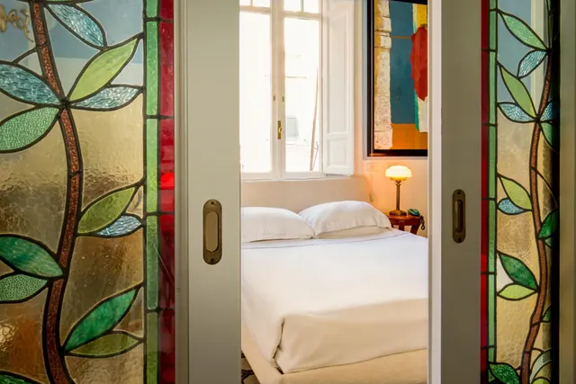 A bed with white linens and a bedside lamp is seen through stained glass sliding doors, with an open window and a painting on the wall.