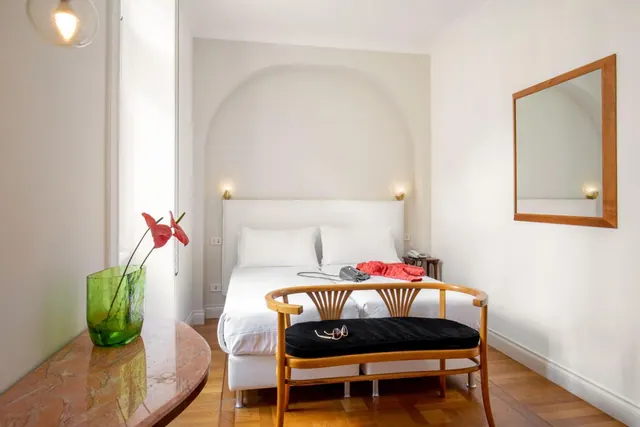 A bedroom with a white double bed in an arched alcove, a wooden bench with a black cushion at the foot of the bed, and a green vase of red flowers on a marble table.