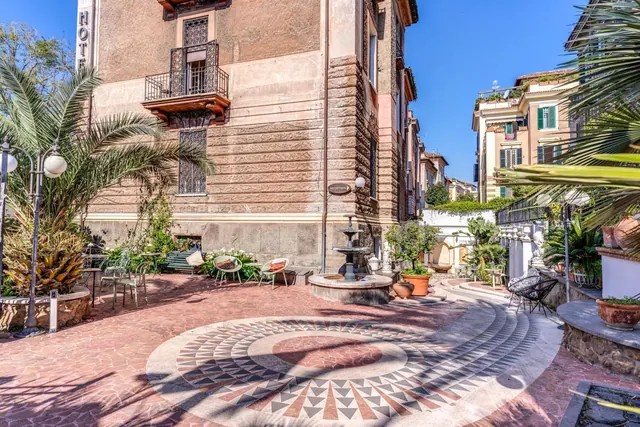 A paved courtyard with a mosaic floor, a stone fountain, palm trees and metal chairs in front of an old multi-storey building.