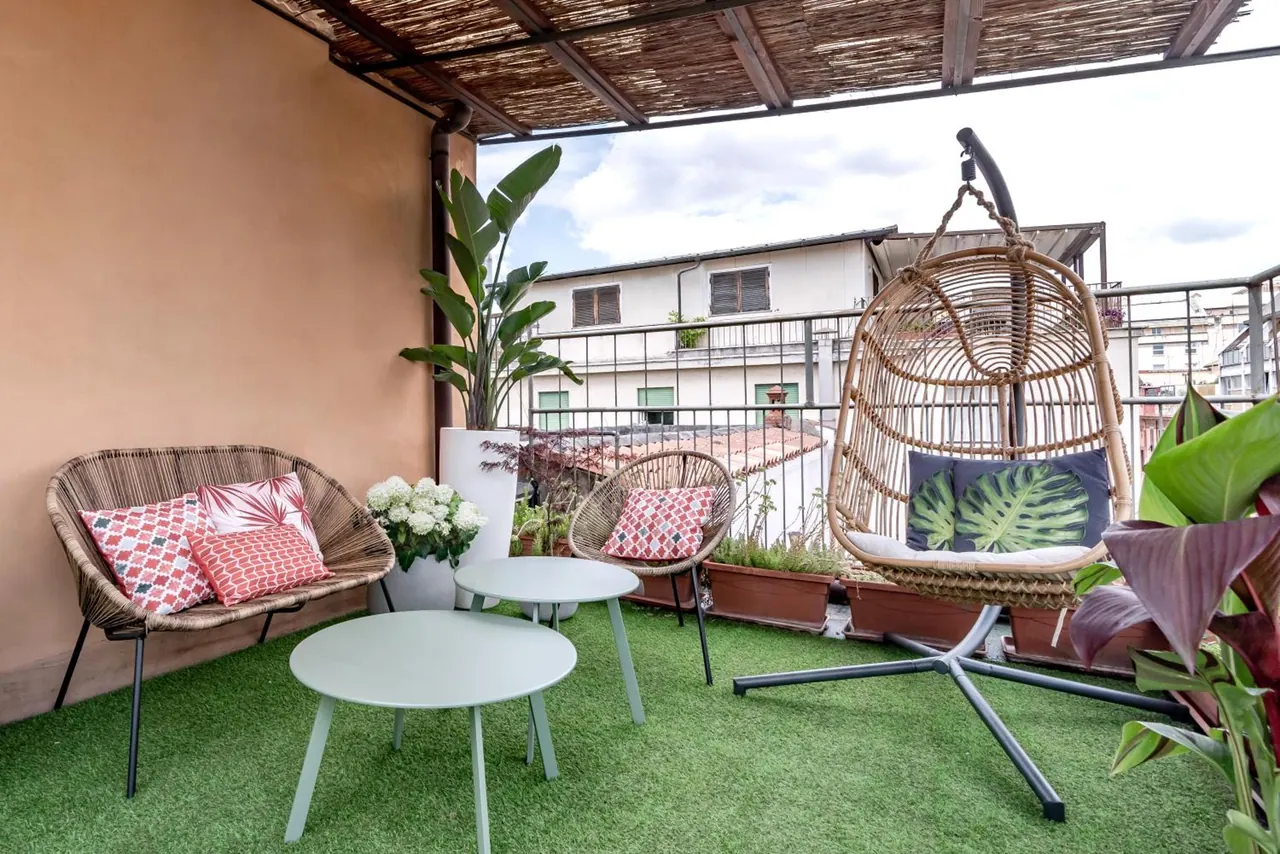 A furnished rooftop terrace with wicker seating, a hanging egg chair, round side tables and artificial grass, with neighbouring buildings visible beyond the railing.