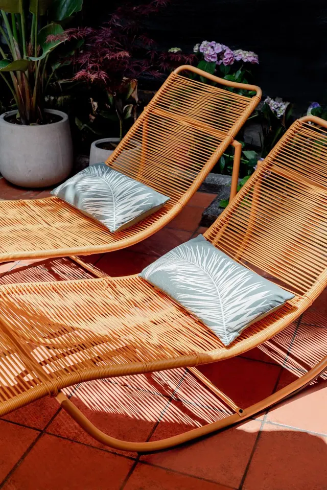 Two orange woven sun loungers with palm-print cushions sit on a terracotta tiled patio beside potted plants and hydrangeas.