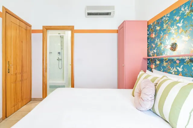 A bedroom with a made white double bed, striped and round cushions, a floral blue wallpapered headboard wall, a pink wardrobe, a wall-mounted air conditioner and an open doorway to a shower.