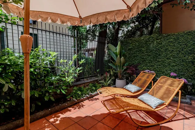 A terracotta-tiled terrace holds two woven rattan lounge chairs with cushions, a beige scalloped parasol, potted plants, and a tall clipped hedge behind.