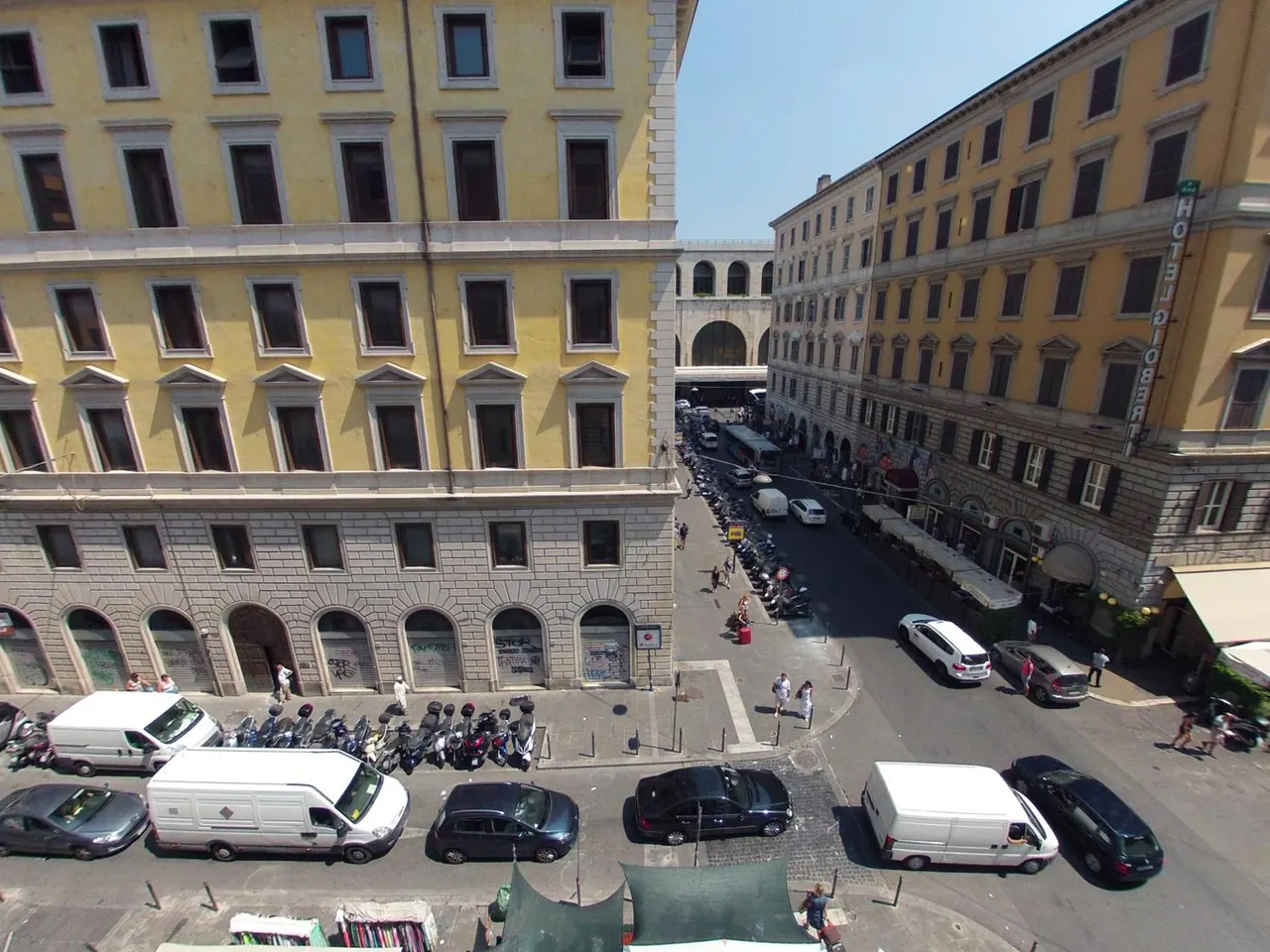 View from an upper-floor window down onto a street lined with parked cars and scooters, with yellow and grey historic buildings on both sides and a vertical hotel sign on the right.