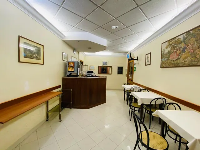 A small dining room with white-clothed tables, cane-back chairs, framed pictures on the walls and a coffee machine on a wooden bar counter.